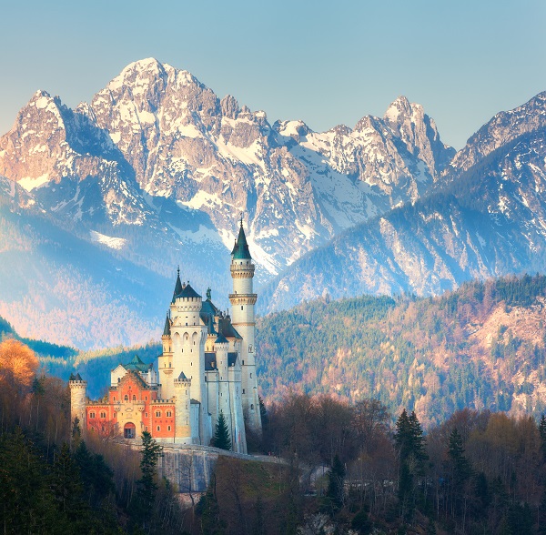 The famous Neuschwanstein Castle in the background of snowy mountains at sunrise in Germany. Beautiful spring landscape with castle, mountain peaks and green trees. Landmarks in Europe. Travel